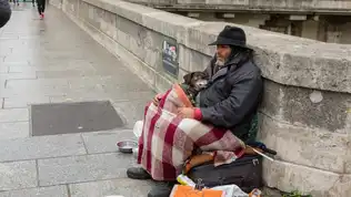 Un homme sans abri assis sur un trottoir à Paris, enveloppé dans une couverture, tenant un petit chien.