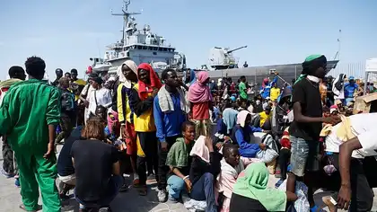 Un groupe de migrants assis et debout sur un quai, avec un navire de garde-côtes en arrière-plan