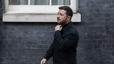 Volodymyr Zelensky marchant devant un mur de briques grises, ajustant le col de sa veste noire.