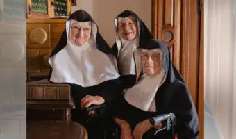 Trois religieuses âgées en habit de congrégation souriant dans un intérieur boisé à Salzbourg.