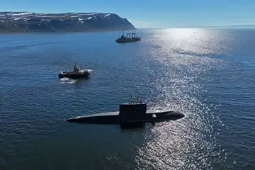 Un sous-marin noir naviguant en surface escorté par un petit navire dans un fjord.