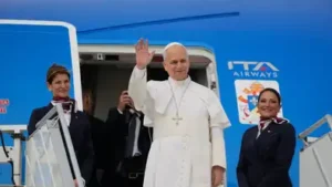 Le Pape Léon XIV saluant de la main à la porte d'un avion entouré d'hôtesses de l'air.
