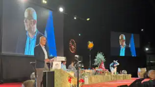 Homme à lunettes parlant au pupitre devant des ballots de paille et l'écran géant d'un congrès.