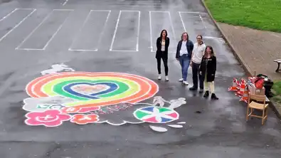 Quatre adultes posant dans une cour de récréation bitumée à côté d'un dessin coloré au sol aux symboles LGBT et fleurs.