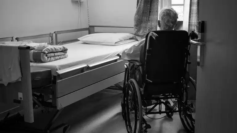 Vue en noir et blanc d'une personne âgée de dos en fauteuil roulant dans une chambre d'hôpital.
