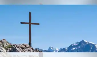 Grande croix latine en bois dressée sur un sommet rocheux face à des montagnes enneigées.
