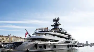 Grand yacht de luxe amarré dans un port urbain ensoleillé sous un ciel bleu parsemé de nuages légers.
